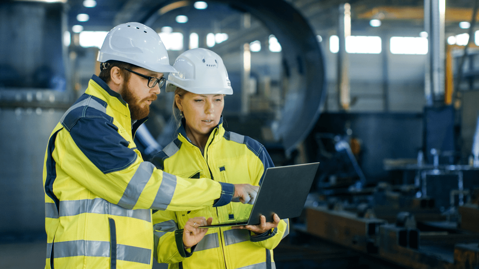 Man and woman working in construction in hi vis and hard hats looking at a laptop